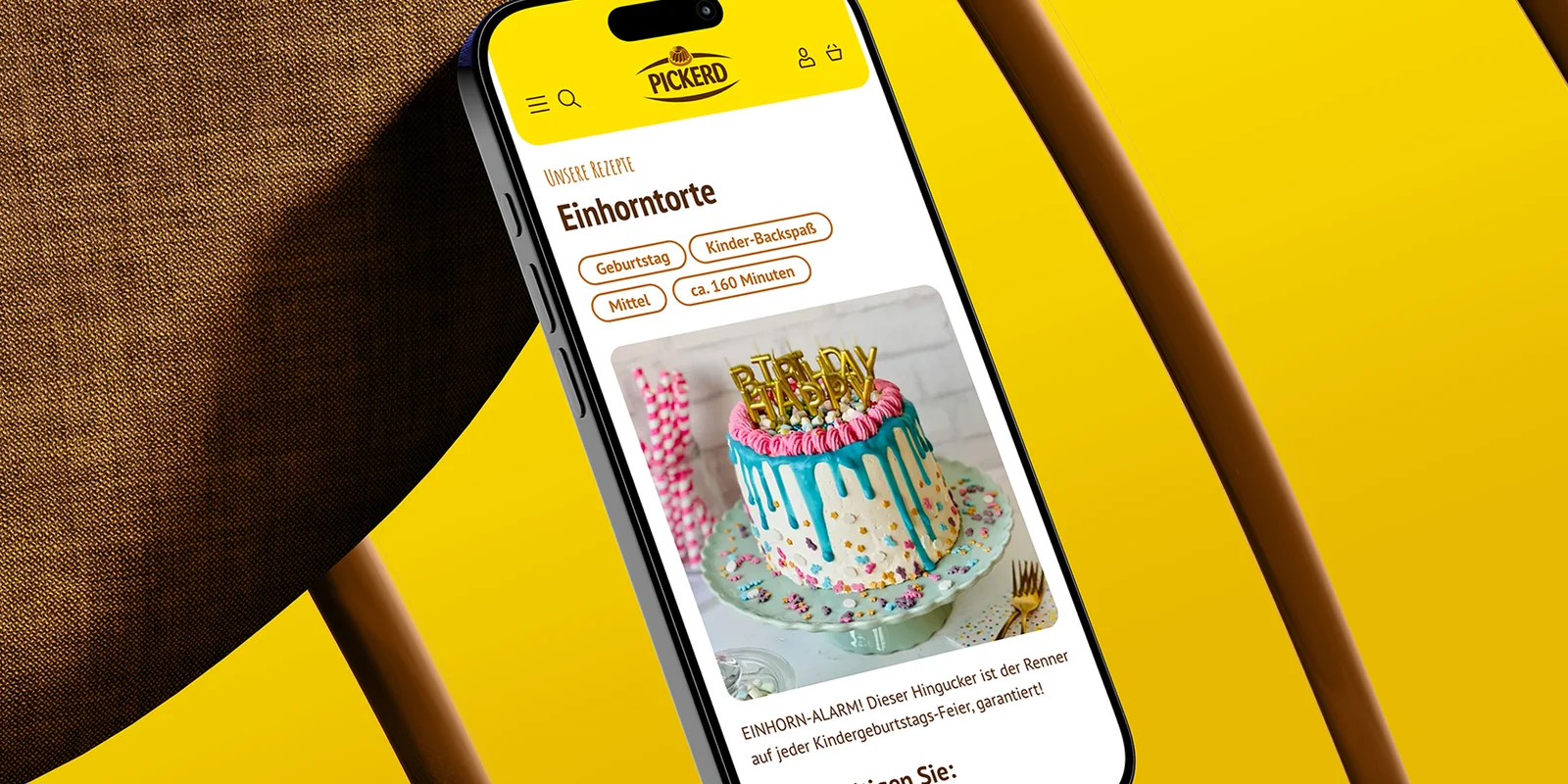 Smartphone vor braun, gelbem Hintergrund mit geöffneter Pickerd-Website zeigt Rezeptbild für eine Happy Birthday-Torte